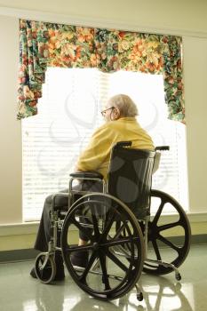 Elderly man in wheelchair sits and looks out of large window. Vertical shot.