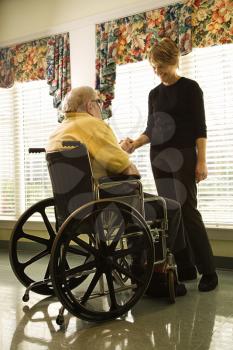Elderly man in a wheelchair and a young woman stand by a window and hold hands.  Vertical shot.