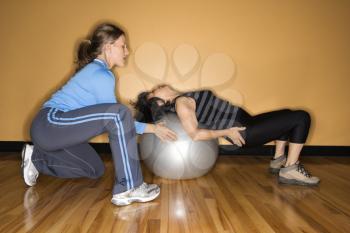 Woman steadies a balance ball for another woman lying back on it at the gym. Horizontal shot.