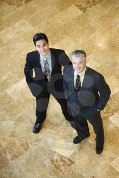 Two business men stand on a marble floor with hands in their pockets. They are looking up towards the camera. Vertical shot.
