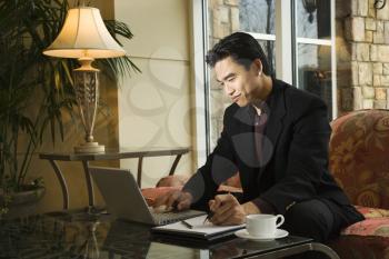 A young Asian businessman typing on a laptop computer whiles taking notes on a notebook. Horizontal shot.