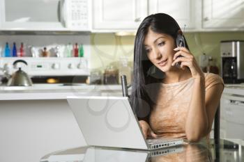 A young woman sits at the kitchen table using a laptop and talking on a cell phone. Horizontal shot.
