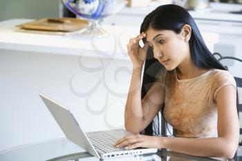 A young woman sitting at the kitchen table using a laptop. She has a worried expression on her face. Horizontal shot.