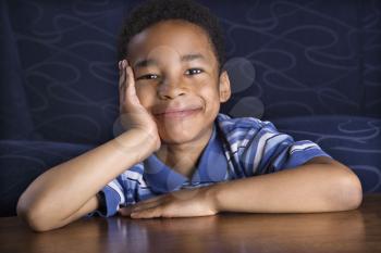 Portrait of a young African American boy sitting in front of a couch with his face propped on his hand. Horizontal shot.