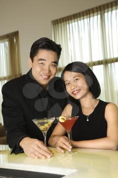 Attractive young Asian couple smiling towards the camera and holding cocktails at a bar. Vertical shot.
