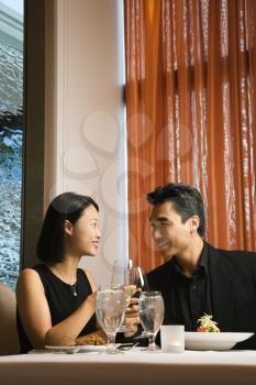 Attractive young Asian couple sit at a restaurant table smiling and toasting their wine. Vertical shot.