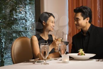 Attractive young Asian couple sit at a restaurant table smiling and toasting. Horizontal shot.