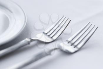 Two forks sitting side-by-side on a dining table with white tablecloth. Horizontal shot.
