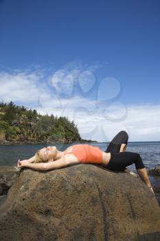 Attractive young woman lies on her back on top of a large rock at the beach. She has her eyes closed. Vertical shot.