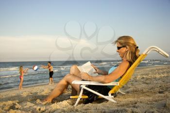 Woman at the beach reads a book while her family plays in the background by the ocean.