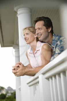 Smiling man and woman look out from a house porch leaning on the railing. Vertical shot.