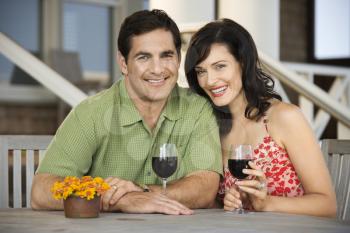 Man and woman smile towards the camera at an outdoor cafe holding glasses of wine. Horizontal shot.
