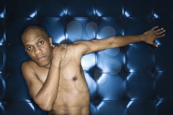 Handsome young man sitting with his hand on his shoulder against a blue padded background. He is shirtless and has one arm outstreatched. Horizontal shot.