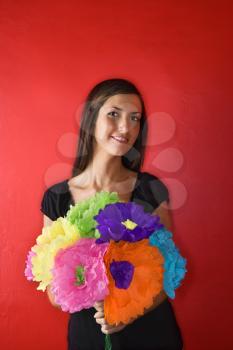Attractive young adult woman standing against a red wall holding a bouquet of colorful paper flowers. Vertical shot. Isolated on red.
