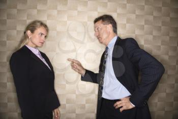 Caucasian middle-aged businessman pointing to and reprimanding mid-adult Caucasian businesswoman. Horizontal format.