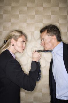 Caucasian mid-adult businesswoman staring into eyes of a middle-aged businessman while pulling angrily on his tie. Vertical format.