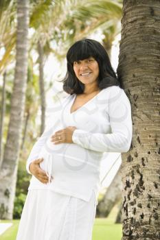 Portrait of a pregnant woman leaning against a tree and holding her belly. Vertical shot.