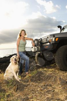 A smiling woman leans against her SUV and stands next to her dog as both look at the camera at a beach. Vertical format.