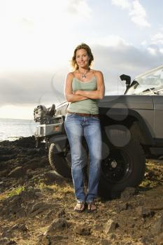 A woman leans against her SUV and smiles at the camera at a beach. Vertical format.
