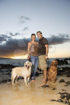 A man and woman smile at the camera as they hold the leashes of their dogs on a beach. Vertical format.