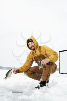 Young man pulls a fish out of a hole in the ice. He is holding a fishing rod and wearing snow gear. Vertical shot.