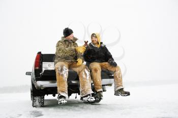 Two young men enjoy a beer while sitting on the tailgate of a truck in a winter environment. Horizontal shot.