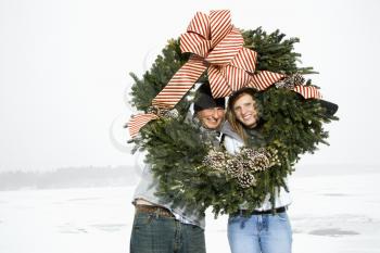 Young adult couple in the winter holding a Christmas wreath. Their heads are framed by the wreath. Horizontal shot.