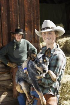 Attractive young woman wearing a cowboy hat and holding an Australian Shepherd. A young man is standing in the background. Vertical shot.
