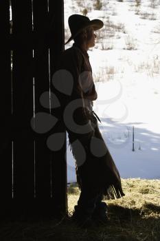 Silhouette of a cowboy leaning in a barn doorway, looking outside. Vertical shot.