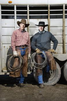 Two men wearing cowboy hats and leaning on the side of a livestock trailer. They are holding lariats. Vertical shot.