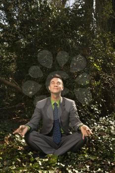 Young businessman sits in a lotus position meditating in the woods with closed eyes and a smile. Vertical shot.
