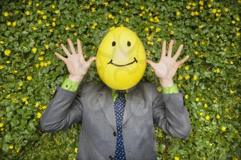 Businessman lies in a flower bed with a yellow balloon happy face on his head. Horizontal shot.