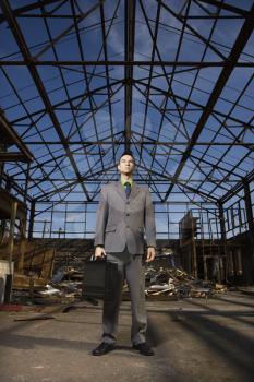 Young businessman stands in an abandoned building holding a briefcase with a serious look on his face. Vertical shot