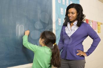 Teacher watching young female student write on blackboard. Horizontally framed shot.