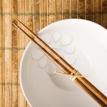 Overhead view of chopsticks lying across an empty bowl on a bamboo mat. Square format.
