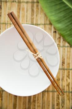 Overhead view of chopsticks lying across an empty bowl on a bamboo mat. Part of a green leaf can be seen in the background. Vertical shot.