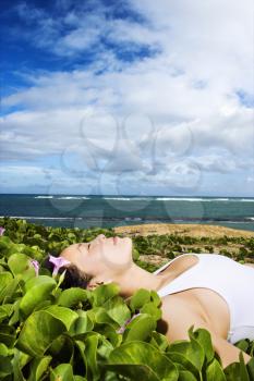 Asian woman lying in a patch of flowers and foliage near a Maui Hawaii beach with the ocean in the background. Vertical shot.