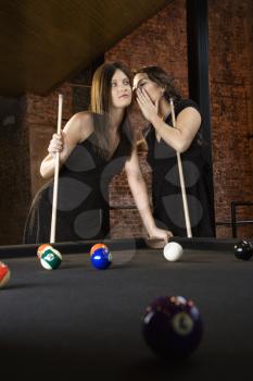 Two attractive young women at pool table as one whispers into other's ear.