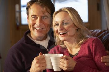 Middle Aged Couple Sitting On Sofa With Hot Drinks Watching TV