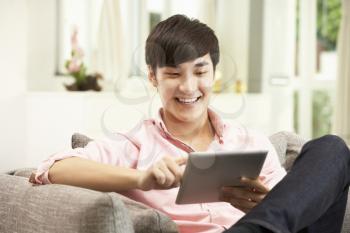 Young Chinese Man Using Digital Tablet Whilst Relaxing On Sofa At Home