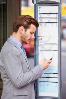Businessman At Bus Stop With Mobile Phone Reading Timetable