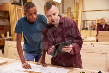 Carpenter With Apprentice Looking At Plans In Workshop