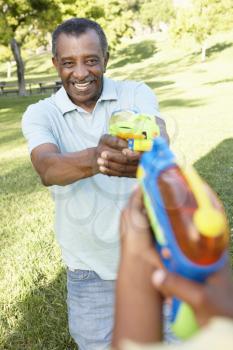 African American Grandfather And Grandson Playing With Water Pistols In Park