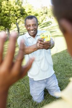 African American Grandfather And Grandson Playing With Water Pistols In Park