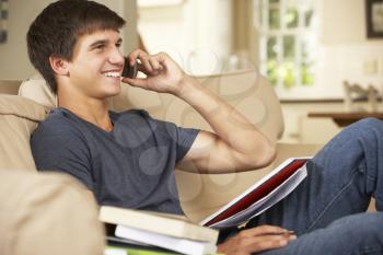 Teenage Boy Sitting On Sofa At Home Doing Homework Using Mobile Phone