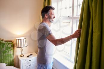Middle aged man opens curtains in hotel room in the morning