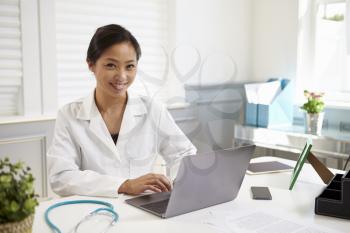 Female Doctor Sitting At Desk Working At Laptop In Office