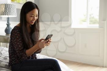 Woman Sitting On Bed In Bedroom Checking Mobile Phone
