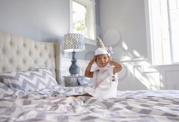 Girl Dressed In Unicorn Costume Sitting On Bed At Home