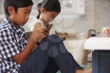 Asian Children Playing Games On Mobile Devices In Kitchen